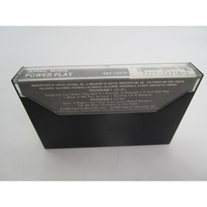 May include: A black cassette tape with a white label. The label has the text "APRIL WINE POWER PLAY" and "4XT-12218" in the top left corner. The label also has the text "7777-12218-4" in the top right corner. The label lists the songs "Anything You Want", "You Got It", "Enough Is Enough", "If You See Kay", "What If We Fall In Love", "Waiting On A Miracle", "Doin' It Right", "Ain't Got Your Love", "Blood Money", "Tell Me Why", "Runners In The Night".