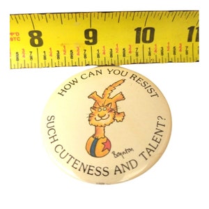 Op de afbeelding: Een ronde knop met een cartoonkat die op een rode en gele bal balanceert. De tekst op de knop luidt "Hoe kun je zo'n schattigheid en talent weerstaan?" met de handtekening "Boynton" onder de bal.