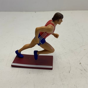 Può includere: Una statuetta di plastica di un corridore in corsa, con un abbigliamento rosso e blu, in piedi su un blocco di partenza in plastica marrone e bianco.