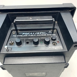 Puede incluir: Sistema de dirección pública inalámbrica FM Realistic negro con una manija en la parte superior. El panel frontal tiene perillas para línea, micrófono y audio inalámbrico. El texto "DC 12.6V 1.2A(MAX)" está impreso en el panel frontal.