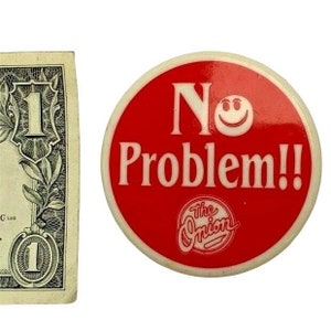 以下が含まれることがあります： 1ドルの紙幣と、赤い丸いボタン。ボタンには白い文字で「No Problem!!」、笑顔の顔、そして「The Onion」というテキストがスタイリッシュなフォントで表示されています。