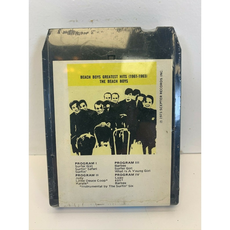 May include: A black cassette tape with a yellow label featuring a photo of The Beach Boys. The label reads "Beach Boys Greatest Hits (1961-1963) The Beach Boys". The back of the cassette tape lists the songs on each side.