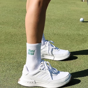May include: White athletic socks with green text that reads "FORE EVER" on the side. The socks are worn with white athletic shoes on a green grassy surface.