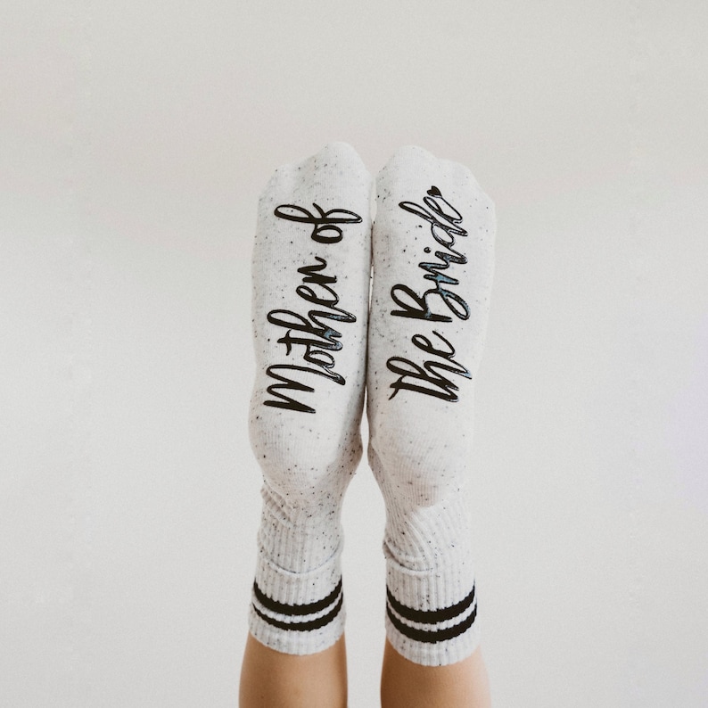 May include: A pair of white socks with black stripes. One sock says "Mother of the Bride" and the other says "The Bride".