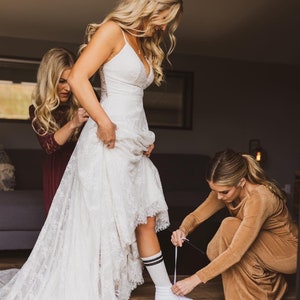 May include: A bride in a white lace wedding dress with a long train is getting ready for her wedding. She is wearing white sneakers with black and white striped socks. Two bridesmaids are helping her put on her shoes.
