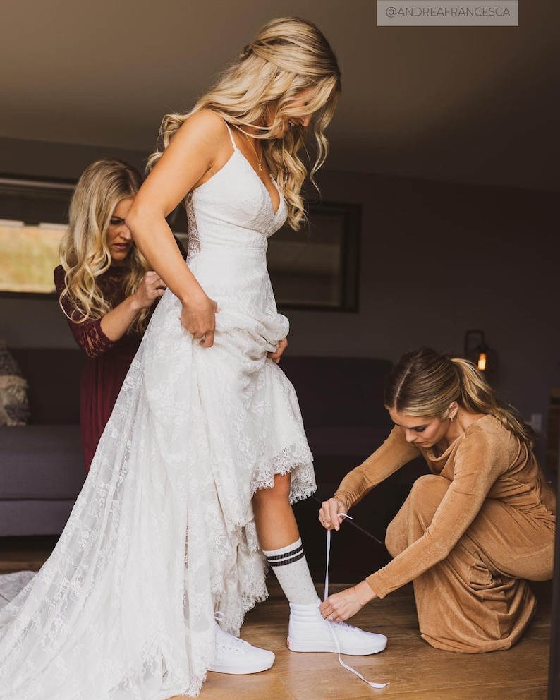 May include: A bride in a white lace wedding dress with a long train is getting ready for her wedding. She is wearing white sneakers with black and white striped socks. Two bridesmaids are helping her put on her shoes.