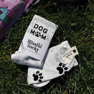Pu&ograve; includere: Un paio di calzini bianchi con impronte di zampe nere e il testo "DOG MOM" su un calzino. I calzini sono su un letto di erba verde. I calzini sono del marchio "Blissful Socks" e hanno il testo "HEELS WITH FEELS" sulla confezione.