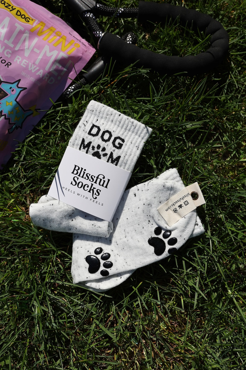 Pu&ograve; includere: Un paio di calzini bianchi con impronte di zampe nere e il testo "DOG MOM" su un calzino. I calzini sono su un letto di erba verde. I calzini sono del marchio "Blissful Socks" e hanno il testo "HEELS WITH FEELS" sulla confezione.