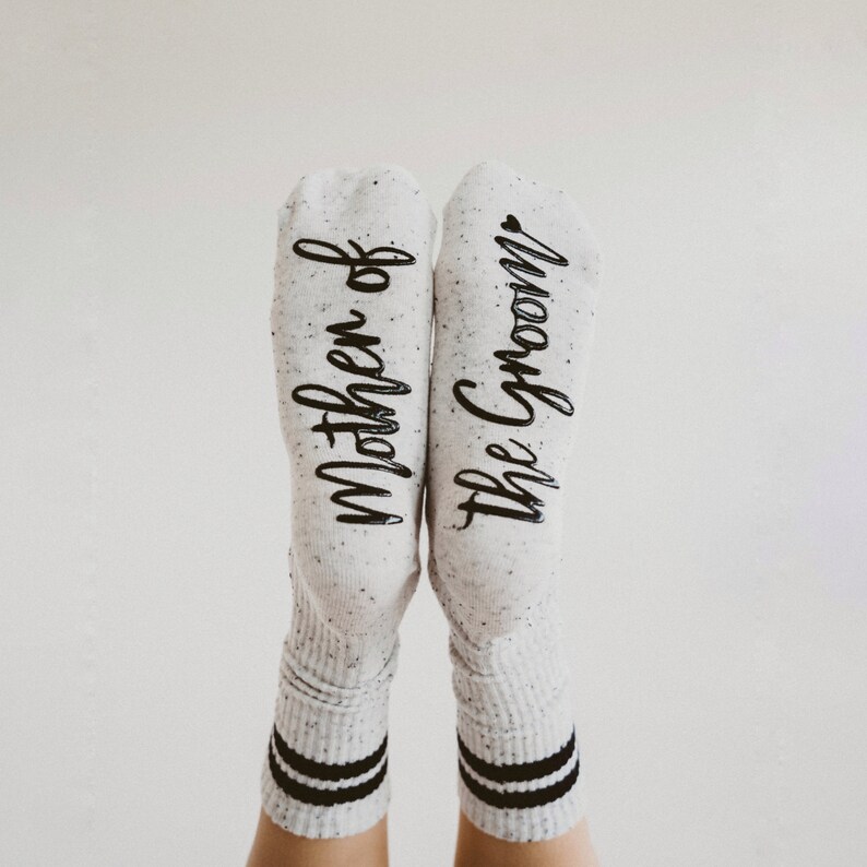 May include: A pair of white socks with black stripes at the top. One sock says "Mother of the Groom" and the other says "Mother of the Groom".