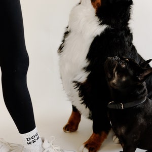 Pu&ograve; includere: Una donna con leggings neri e scarpe da ginnastica bianche con la scritta "DOG MOM" sulle calze, in piedi con due cani. Uno dei cani &egrave; un Bovaro Bernese bianco e nero che guarda in alto, e l'altro &egrave; un Bulldog Francese nero che guarda la fotocamera.
