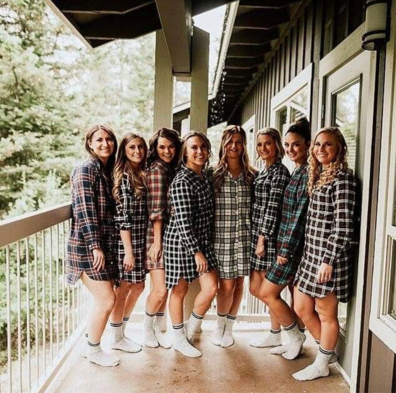 May include: Seven women wearing matching plaid flannel shirts and white socks. The women are standing on a porch with a wooden railing and a view of trees.