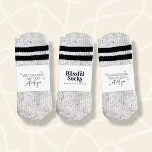 May include: Three pairs of white crew socks with black stripes. The socks have a speckled pattern and are packaged with a white label that says "Blissful Socks" and "Heels with Feels". The labels on the socks say "We couldn't say 'I do' without you", "Our wedding would sock without you", and "Blissful Socks".