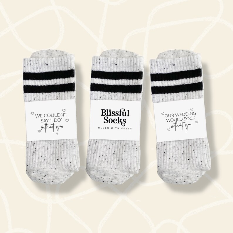 May include: Three pairs of white crew socks with black stripes. The socks have a speckled pattern and are packaged with a white label that says "Blissful Socks" and "Heels with Feels". The labels on the socks say "We couldn't say 'I do' without you", "Our wedding would sock without you", and "Blissful Socks".