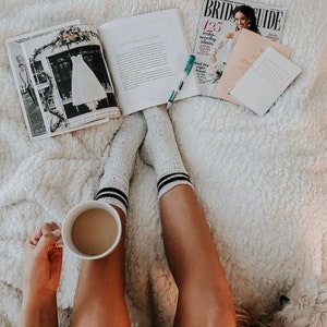 May include: A white fuzzy blanket with a person wearing white socks with black stripes and holding a white coffee mug. The person is reading a magazine about weddings with the text "Bride Guide" and "125 Insta-worthy Ideas".