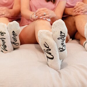 May include: Five pairs of white socks with black lettering. The socks say "The Brides", "Mother of the Bride", "Matron of Honor", "Bridesmaids", and "Mrs. Brides".