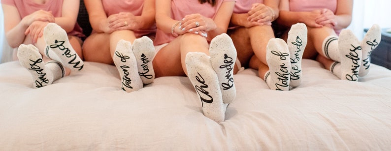 May include: Five pairs of white socks with black lettering. The socks say "The Brides", "Mother of the Bride", "Matron of Honor", "Bridesmaids", and "Mrs. Brides".