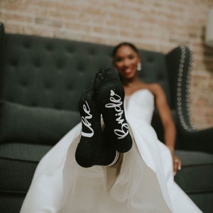 May include: A bride wearing a white wedding dress with black socks that say "the bride" on them. The bride is sitting on a black couch with a brick wall in the background.