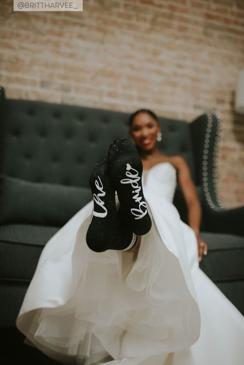 May include: A bride wearing a white wedding dress with black socks that say "the bride" on them. The bride is sitting on a black couch with a brick wall in the background.