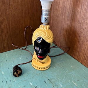 May include: A ceramic table lamp base shaped like a head wearing a yellow turban. The face is black with red lips and a gold accent on the forehead. A silver metal socket holds a white LED light bulb. A brown electrical cord with a plug is attached.