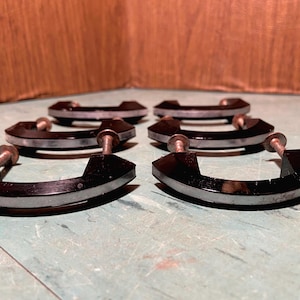 May include: Six vintage cabinet pulls. Each pull is black with a silver accent and a screw post. The pulls are arranged in two rows of three, with the screws facing the viewer. The background is a wooden surface.