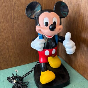 K&ouml;nnte beinhalten: Ein Mickey-Maus-Telefon mit einem K&ouml;rper aus schwarzem, rotem und gelbem Kunststoff. Die Figur steht auf einem schwarzen Sockel, tr&auml;gt wei&szlig;e Handschuhe und h&auml;lt einen blauen H&ouml;rer. Ein schwarzes, gewendeltes Telefonkabel ist am Sockel befestigt.