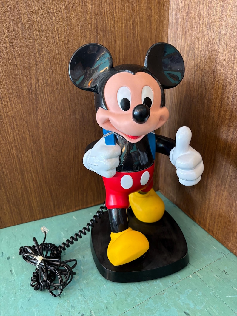 K&ouml;nnte beinhalten: Ein Mickey-Maus-Telefon mit einem K&ouml;rper aus schwarzem, rotem und gelbem Kunststoff. Die Figur steht auf einem schwarzen Sockel, tr&auml;gt wei&szlig;e Handschuhe und h&auml;lt einen blauen H&ouml;rer. Ein schwarzes, gewendeltes Telefonkabel ist am Sockel befestigt.