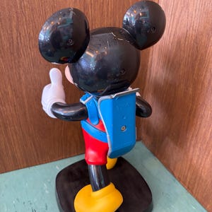 K&ouml;nnte beinhalten: Eine Kunststofffigur von Mickey Mouse in der R&uuml;ckansicht mit gro&szlig;en schwarzen Ohren, einem blauen Rucksack mit dem eingepr&auml;gten Text TYCO, roten Shorts und gelben Schuhen. Die Figur steht auf einem schwarzen Sockel mit einer erhobenen wei&szlig;en Hand.