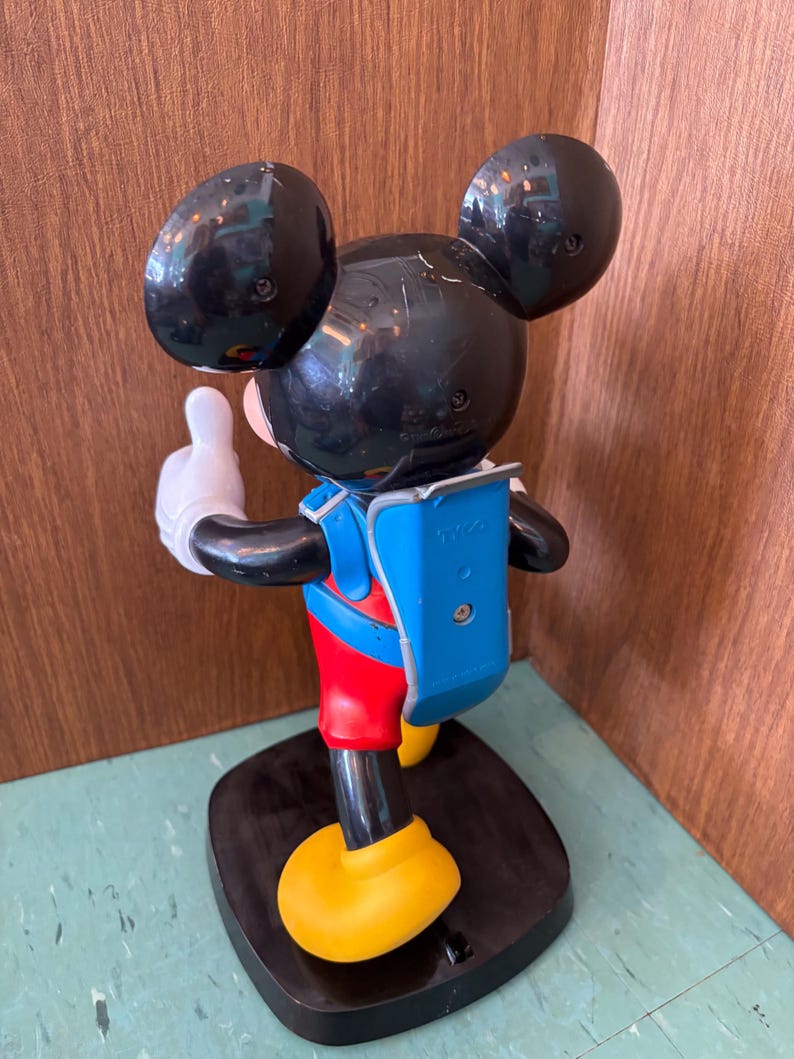 K&ouml;nnte beinhalten: Eine Kunststofffigur von Mickey Mouse in der R&uuml;ckansicht mit gro&szlig;en schwarzen Ohren, einem blauen Rucksack mit dem eingepr&auml;gten Text TYCO, roten Shorts und gelben Schuhen. Die Figur steht auf einem schwarzen Sockel mit einer erhobenen wei&szlig;en Hand.