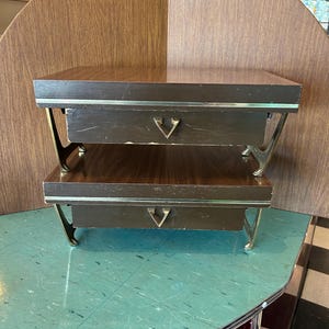 May include: A two-tiered, mid-century modern side table with a dark wood finish. The table has a drawer on each level with a decorative metal pull. The legs are a gold color and have a unique, curved design.