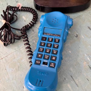 K&ouml;nnte beinhalten: Ein leuchtend blauer Telefonh&ouml;rer mit einem schwarzen Spiralkabel. Das Ger&auml;t verf&uuml;gt &uuml;ber ein Standard-Tastenfeld mit schwarzen Tasten, eine Stummschalttaste, eine Wahlwiederholungstaste und einen Impuls-Ton-Schalter. Das Wort Tyco ist unten eingepr&auml;gt.