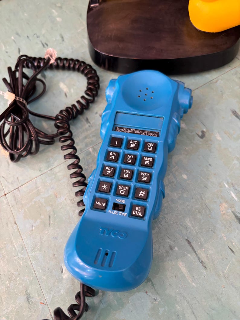 K&ouml;nnte beinhalten: Ein leuchtend blauer Telefonh&ouml;rer mit einem schwarzen Spiralkabel. Das Ger&auml;t verf&uuml;gt &uuml;ber ein Standard-Tastenfeld mit schwarzen Tasten, eine Stummschalttaste, eine Wahlwiederholungstaste und einen Impuls-Ton-Schalter. Das Wort Tyco ist unten eingepr&auml;gt.