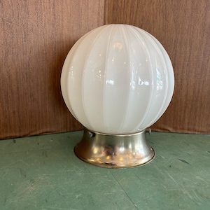 May include: A white, ribbed glass globe lamp with a brass-coloured metal base. The globe has a round shape and is mounted on a circular base. The lamp is designed for indoor use and provides ambient lighting.