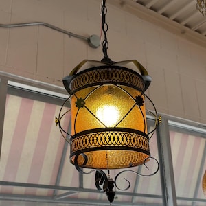 May include: A vintage hanging lantern with a warm amber glass shade. The lantern features ornate black metal filigree and a decorative tassel. The light fixture is suspended from a chain and is likely for indoor use.