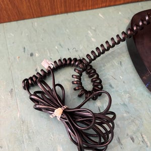 K&ouml;nnte beinhalten: Ein schwarzes, spiralf&ouml;rmiges Telefonkabel mit einem transparenten Kunststoff-Stecker an einem Ende. Das Kabel ist mit einem kleinen, hellen Kunststoff-Kabelbinder zusammengebunden, wodurch Schlaufen und eine eng gewickelte Spirale entstehen.