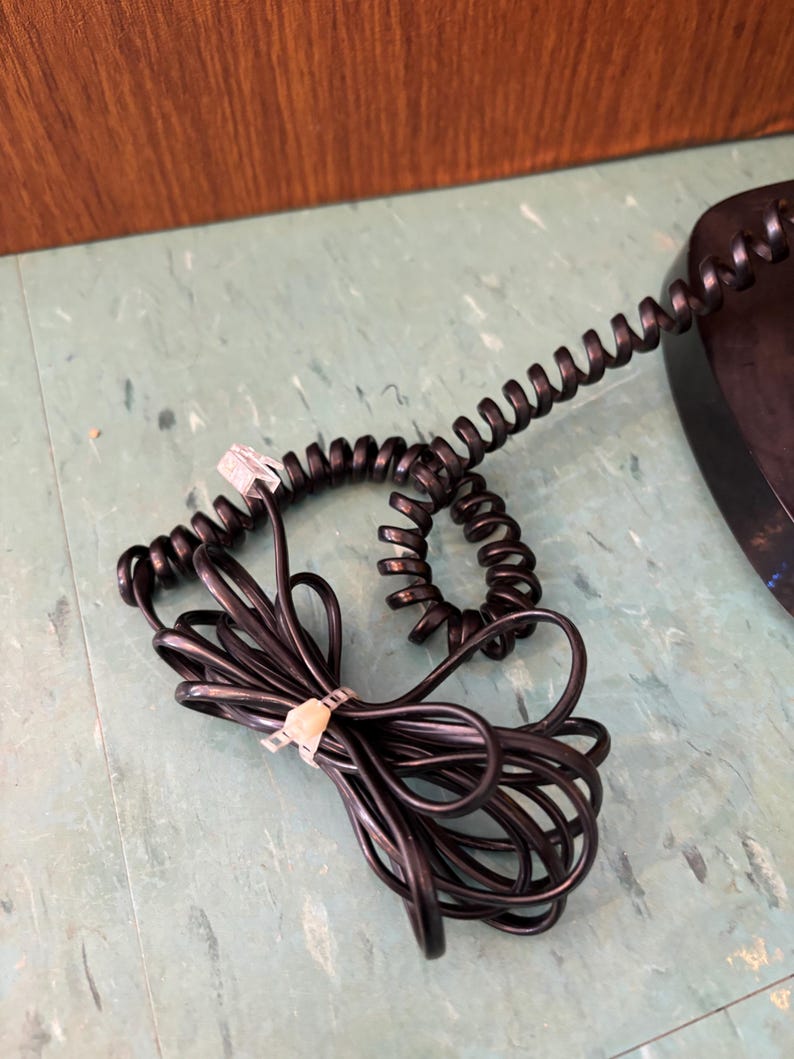 K&ouml;nnte beinhalten: Ein schwarzes, spiralf&ouml;rmiges Telefonkabel mit einem transparenten Kunststoff-Stecker an einem Ende. Das Kabel ist mit einem kleinen, hellen Kunststoff-Kabelbinder zusammengebunden, wodurch Schlaufen und eine eng gewickelte Spirale entstehen.