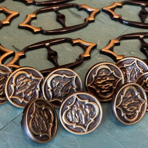 May include: A collection of antique brass hardware, including drawer pulls and decorative knobs. The drawer pulls are black with gold accents, and the knobs feature a textured design within a gold frame. The hardware is arranged on a green surface.