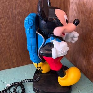 K&ouml;nnte beinhalten: Ein Mickey-Maus-Telefon in den Farben Schwarz, Rot und Gelb. Die Figur tr&auml;gt einen blauen Rucksack und steht auf einem schwarzen Sockel. Ein schwarzes, spiralf&ouml;rmiges Telefonkabel f&uuml;hrt vom Rucksack zur Basisstation des Ger&auml;ts.