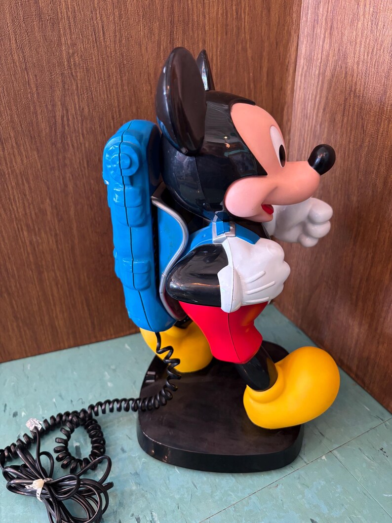 K&ouml;nnte beinhalten: Ein Mickey-Maus-Telefon in den Farben Schwarz, Rot und Gelb. Die Figur tr&auml;gt einen blauen Rucksack und steht auf einem schwarzen Sockel. Ein schwarzes, spiralf&ouml;rmiges Telefonkabel f&uuml;hrt vom Rucksack zur Basisstation des Ger&auml;ts.