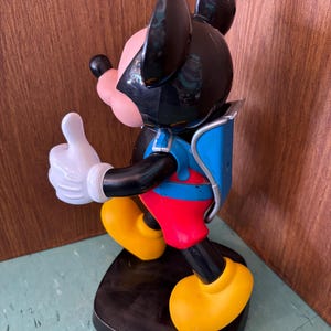 K&ouml;nnte beinhalten: Eine Mickey-Maus-Figur auf einem schwarzen Sockel mit schwarzem Kopf und gro&szlig;en Ohren, einem rosa Gesicht und einer wei&szlig;en Handschuhhand, die einen Daumen nach oben zeigt. Die Figur tr&auml;gt eine rote Hose, einen blauen Rucksack und gelbe Schuhe.