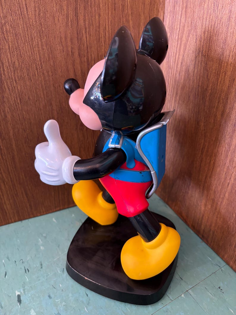 K&ouml;nnte beinhalten: Eine Mickey-Maus-Figur auf einem schwarzen Sockel mit schwarzem Kopf und gro&szlig;en Ohren, einem rosa Gesicht und einer wei&szlig;en Handschuhhand, die einen Daumen nach oben zeigt. Die Figur tr&auml;gt eine rote Hose, einen blauen Rucksack und gelbe Schuhe.