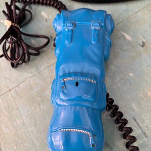 K&ouml;nnte beinhalten: Ein leuchtend blauer Telefonh&ouml;rer in Form eines Rucksacks. Der H&ouml;rer weist geformte Details wie Riemen, Schnallen und Rei&szlig;verschlusstaschen auf. Ein langes, schwarzes Spiralkabel ist oben und unten an der blauen Kunststoffeinheit befestigt.
