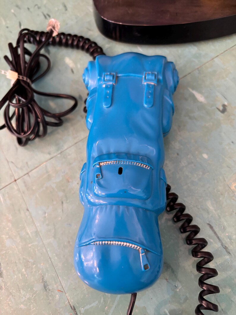 K&ouml;nnte beinhalten: Ein leuchtend blauer Telefonh&ouml;rer in Form eines Rucksacks. Der H&ouml;rer weist geformte Details wie Riemen, Schnallen und Rei&szlig;verschlusstaschen auf. Ein langes, schwarzes Spiralkabel ist oben und unten an der blauen Kunststoffeinheit befestigt.