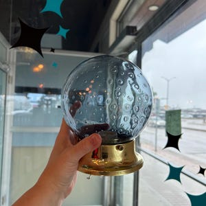 May include: A spherical blue glass light fixture shade featuring a textured bubble pattern throughout the surface. The glass globe is attached to a circular, polished gold-tone metal base with a small screw visible on the side of the fixture.