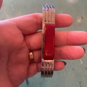 Peut inclure: Un bracelet argenté avec une pierre centrale rectangulaire rouge foncé. Le bracelet présente un motif nervuré sur les côtés. Le bracelet est présenté sur une main.