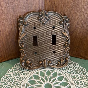 May include: Ornate bronze-colored double light switch cover with a diamond pattern and decorative scrollwork. The cover has two rectangular switch openings and two screw holes. The background is a wooden surface.