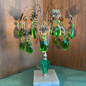 May include: A vintage table lamp with a silver metal frame, gold-toned floral details, and hanging green crystal droplets. The lamp has a green glass base and rests on a white marble square base. The lamp is plugged in.