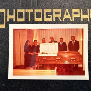 May include: A vintage photograph in a black album with gold lettering that reads "PHOTOGRAPHS." The photo shows a casket with people standing around it. The people are dressed in dark clothing. The casket is made of wood.