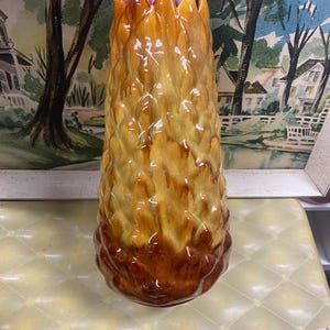 May include: A tall, textured ceramic vase with a pineapple-like design. The vase features a gradient of colors, transitioning from a dark brown base to shades of yellow and orange at the top. The vase is held by a hand.