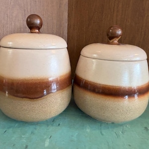 May include: Two ceramic jars with lids. The jars are beige with a brown stripe around the middle and a brown knob on top. The jars are on a green surface.
