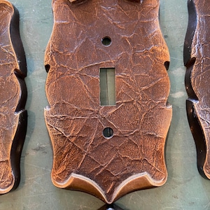 May include: Decorative brown and bronze light switch cover with a textured leather-like appearance. The cover has a shield-like shape with a toggle switch opening and two screw holes. The edges are bronze-coloured.